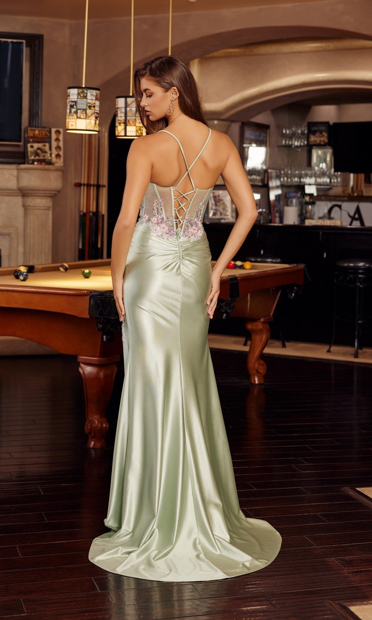 A woman wearing the Nox Anabel G1789 long prom dress in light green with a lace-up back stands by a pool table in a room featuring dark wood floors and a bar.